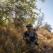 A U.S. Army soldier in full Operational Camouflage Pattern (OCP) uniform is kneeling in a dry, brushy terrain while holding an assault rifle. The soldier is wearing a boonie hat, sunglasses, and a rucksack, suggesting a field training exercise or patrol operation. The environment appears to be arid scrubland with dry grass and sparse trees under a clear sky.