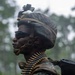 A U.S. Marine in full combat gear is photographed in profile against a blurred green woodland background. The Marine is wearing MARPAT digital camouflage utilities and helmet with a mounted device, dark tactical sunglasses, and carries a belt of linked ammunition draped around the neck and torso. The image is a close-up portrait-style photograph taken in what appears to be a rainy outdoor training or operational environment.