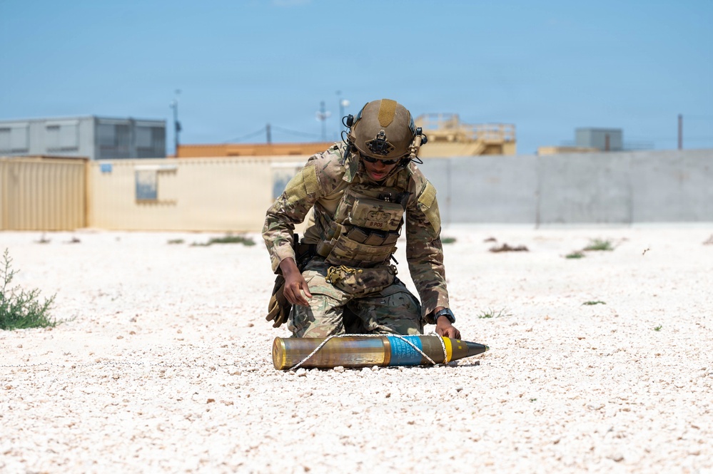 A military service member in full combat gear is kneeling on a gravel surface, handling what appears to be an artillery round or large munition with blue and yellow markings. The setting appears to be a forward operating base or military installation in an arid environment. The activity suggests ordnance handling, training, or an explosive ordnance disposal (EOD) exercise.