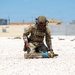 A military service member in full combat gear is kneeling on a gravel surface, handling what appears to be an artillery round or large munition with blue and yellow markings. The setting appears to be a forward operating base or military installation in an arid environment. The activity suggests ordnance handling, training, or an explosive ordnance disposal (EOD) exercise.