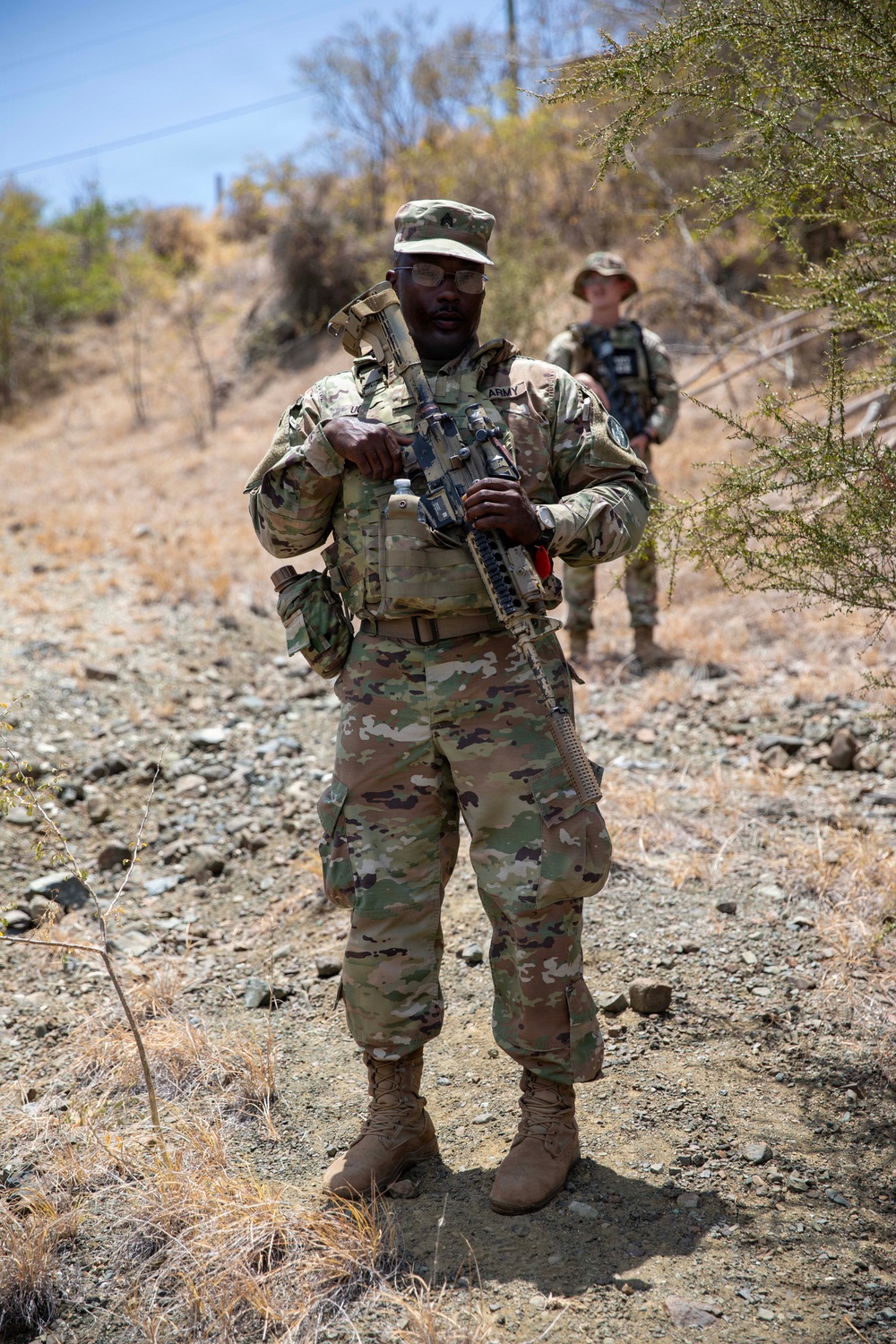 Two U.S. Army soldiers in Operational Camouflage Pattern (OCP) uniforms are standing on a rocky, arid terrain with dry vegetation in the background. The foreground soldier is holding an assault rifle at a relaxed ready position and wearing glasses, with 'ARMY' tape visible on the chest. The environment suggests an overseas deployment or training exercise in an arid/semi-arid region.