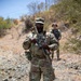 Two U.S. Army soldiers in Operational Camouflage Pattern (OCP) uniforms are standing on a rocky, arid terrain with dry vegetation in the background. The foreground soldier is holding an assault rifle at a relaxed ready position and wearing glasses, with 'ARMY' tape visible on the chest. The environment suggests an overseas deployment or training exercise in an arid/semi-arid region.