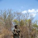 A soldier in full combat gear stands in an arid, scrubland environment with dry grass and sparse trees under a blue sky. The individual is wearing digital camouflage uniform consistent with USMC MARPAT pattern, tactical vest, boonie hat, and sunglasses while holding a rifle. The setting appears to be a field training exercise or patrol in a dry, semi-arid region.