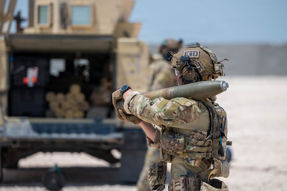 A soldier in full multicam combat gear carries what appears to be an artillery round or large munition over their shoulder in an arid, open environment. The helmet patch visible reads 'EOD' (Explosive Ordnance Disposal), suggesting this individual is part of a bomb disposal unit. A military vehicle, likely a HMMWV (Humvee), is visible in the background along with at least one other personnel.