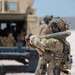 A soldier in full multicam combat gear carries what appears to be an artillery round or large munition over their shoulder in an arid, open environment. The helmet patch visible reads 'EOD' (Explosive Ordnance Disposal), suggesting this individual is part of a bomb disposal unit. A military vehicle, likely a HMMWV (Humvee), is visible in the background along with at least one other personnel.