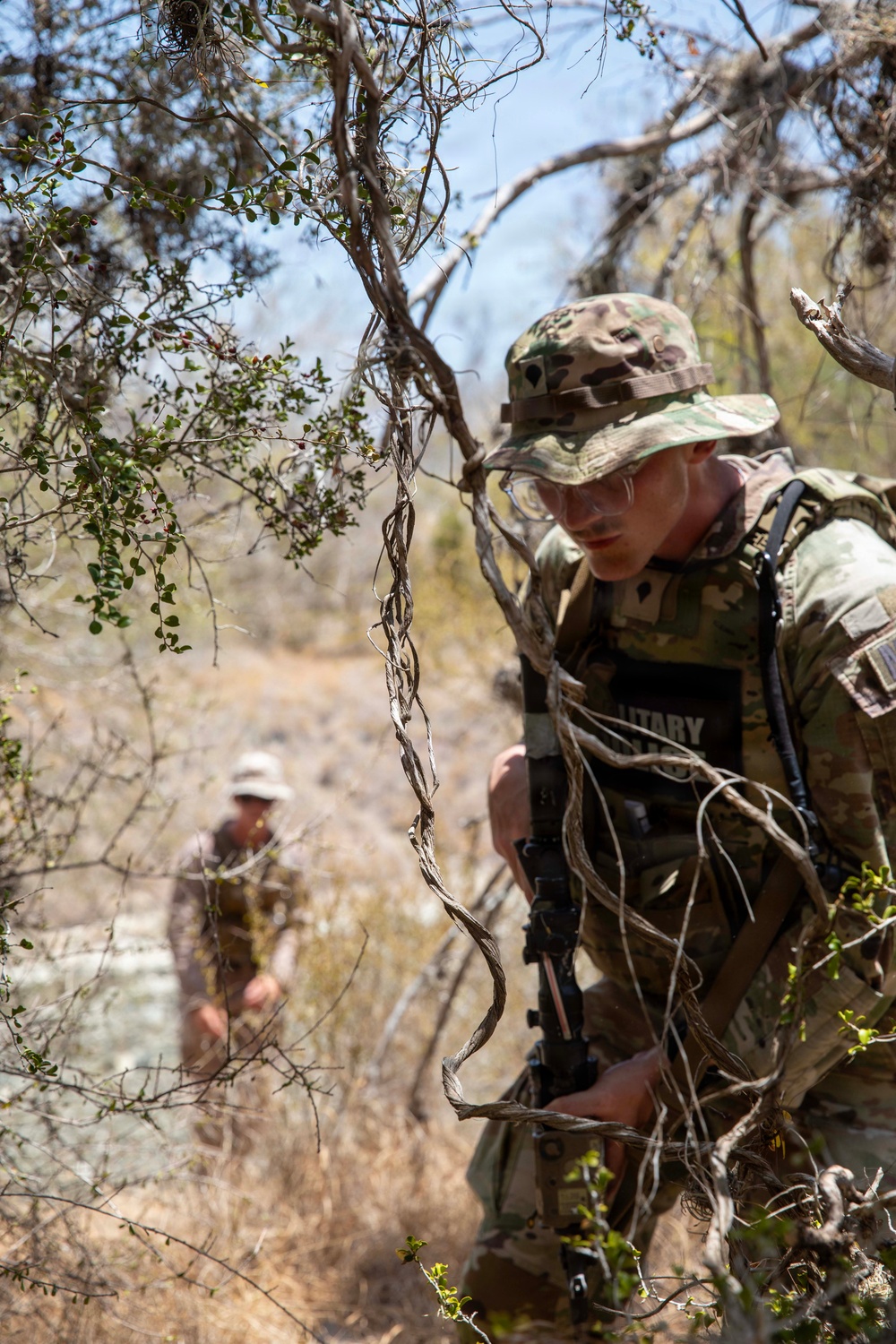 Two military personnel in camouflage uniforms navigate through dense, dry scrubland terrain. The foreground soldier wears a boonie hat and multicam pattern uniform, carries a rifle, and wears a vest with partially visible text reading 'MILITARY'. A second soldier is visible in the background, similarly dressed, in what appears to be a field training exercise.