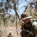 Two military personnel in camouflage uniforms navigate through dense, dry scrubland terrain. The foreground soldier wears a boonie hat and multicam pattern uniform, carries a rifle, and wears a vest with partially visible text reading 'MILITARY'. A second soldier is visible in the background, similarly dressed, in what appears to be a field training exercise.