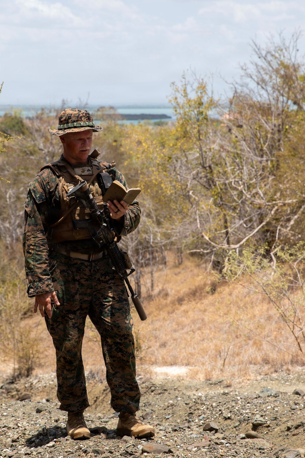 A U.S. Marine in full MARPAT woodland digital camouflage uniform stands on rocky elevated terrain, reviewing what appears to be a notebook or small document. He is equipped with a tactical vest, slung rifle (appears to be an M4 carbine), and a boonie cover. The background shows dry scrub vegetation and a body of water visible in the distance, suggesting a coastal or island environment.