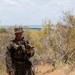 A U.S. Marine in full MARPAT woodland digital camouflage uniform stands on rocky elevated terrain, reviewing what appears to be a notebook or small document. He is equipped with a tactical vest, slung rifle (appears to be an M4 carbine), and a boonie cover. The background shows dry scrub vegetation and a body of water visible in the distance, suggesting a coastal or island environment.