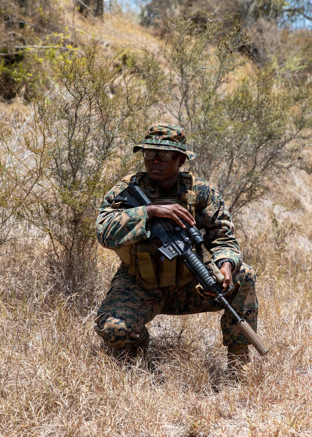 A military service member in MARPAT (Marine pattern) digital camouflage uniform is kneeling in a dry, scrubby outdoor terrain. The individual is holding an assault rifle and wearing tactical gear including a plate carrier vest and a boonie hat. The setting appears to be a field training exercise in an arid or semi-arid environment.