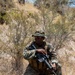 A military service member in MARPAT (Marine pattern) digital camouflage uniform is kneeling in a dry, scrubby outdoor terrain. The individual is holding an assault rifle and wearing tactical gear including a plate carrier vest and a boonie hat. The setting appears to be a field training exercise in an arid or semi-arid environment.
