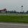 A red aerobatic aircraft (registration N756K) is performing a low-altitude inverted or banked maneuver over an airport runway. Several ground crew members wearing high-visibility yellow vests are positioned along the runway, likely serving as safety marshals or judges. A ribbon or streamer line is strung between a light pole and a stake, possibly serving as a target or marker for the aerobatic performance.