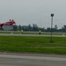 A red aerobatic aircraft (registration N756K) is performing a low-altitude inverted or banked maneuver over an airport runway. Several ground crew members wearing high-visibility yellow vests are positioned along the runway, likely serving as safety marshals or judges. A ribbon or streamer line is strung between a light pole and a stake, possibly serving as a target or marker for the aerobatic performance.