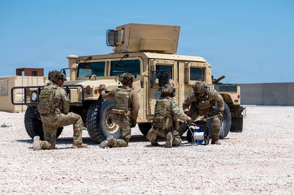 Four military personnel in full combat gear are positioned around a desert-tan HMMWV (Humvee) on a gravel surface in an arid environment. The soldiers appear to be conducting equipment setup or maintenance, with at least one handling what appears to be a spool of wire or cable. The setting suggests a forward operating base or training area in a desert region.