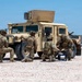 Four military personnel in full combat gear are positioned around a desert-tan HMMWV (Humvee) on a gravel surface in an arid environment. The soldiers appear to be conducting equipment setup or maintenance, with at least one handling what appears to be a spool of wire or cable. The setting suggests a forward operating base or training area in a desert region.