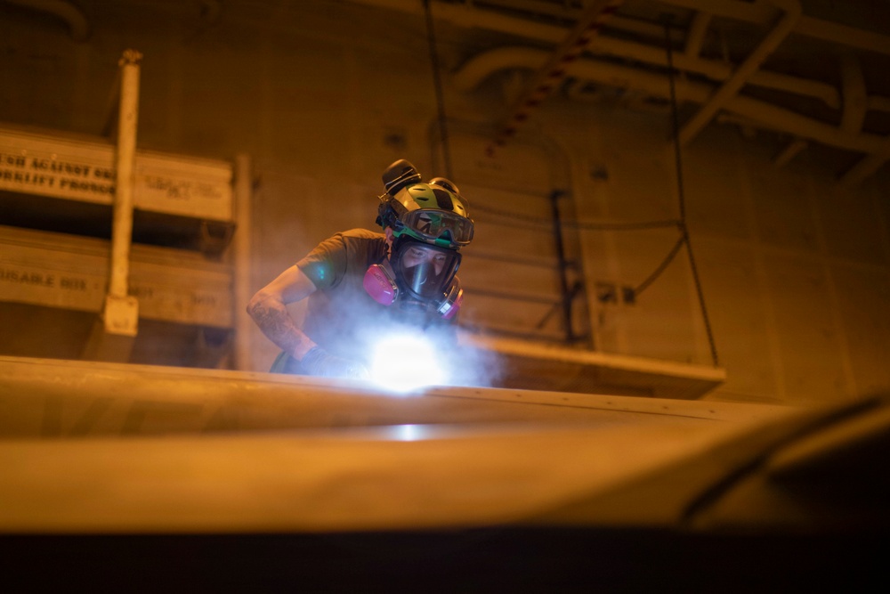 A worker wearing full respiratory protection and safety goggles is performing a grinding or welding operation in what appears to be an industrial or naval vessel interior. A bright light source, likely from the tool being used, illuminates smoke or dust particles in the immediate work area. The environment is dimly lit with an orange ambient glow, suggesting a confined or below-deck workspace.