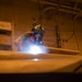 A worker wearing full respiratory protection and safety goggles is performing a grinding or welding operation in what appears to be an industrial or naval vessel interior. A bright light source, likely from the tool being used, illuminates smoke or dust particles in the immediate work area. The environment is dimly lit with an orange ambient glow, suggesting a confined or below-deck workspace.