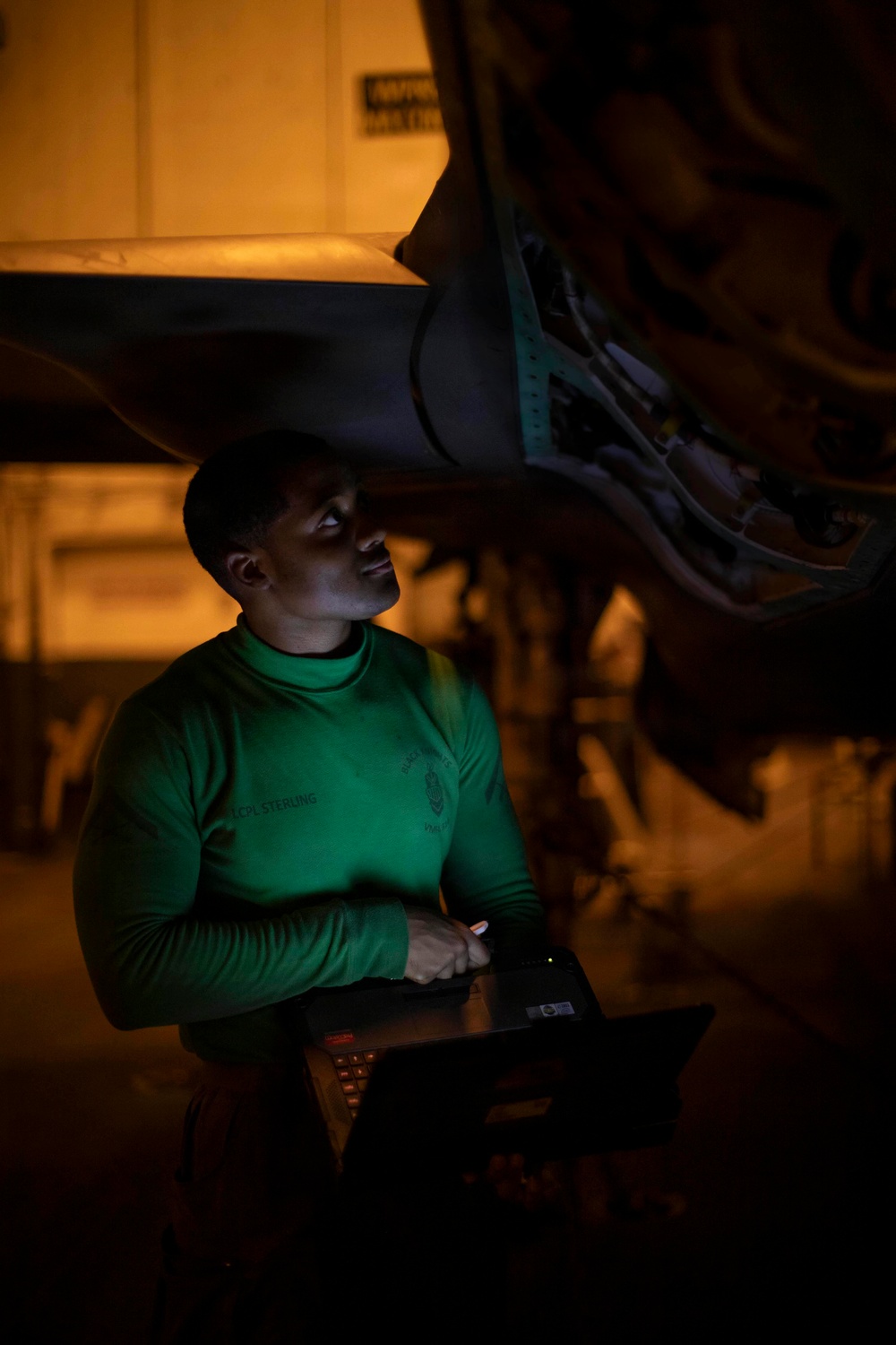A person wearing a green long-sleeve shirt labeled 'LCPL STERLING' holds a laptop computer while looking upward at the underside of what appears to be a military aircraft. The scene is photographed in low-light conditions, with the laptop screen providing green-tinted illumination on the subject's face and torso. The setting appears to be a hangar or flight deck during nighttime operations.