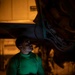 A person wearing a green long-sleeve shirt labeled 'LCPL STERLING' holds a laptop computer while looking upward at the underside of what appears to be a military aircraft. The scene is photographed in low-light conditions, with the laptop screen providing green-tinted illumination on the subject's face and torso. The setting appears to be a hangar or flight deck during nighttime operations.