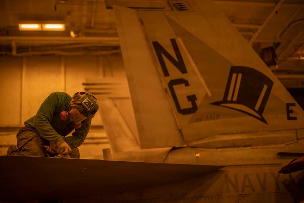 A Navy aircraft maintainer wearing a green shirt, helmet marked '2M', and ear protection is performing maintenance work on an aircraft in what appears to be a carrier hangar bay. The aircraft's tail fin displays the tail code 'NG' and a top hat insignia with 'est. 1919', identifying it as belonging to a specific naval aviation squadron. The scene is illuminated by warm amber/orange hangar lighting.