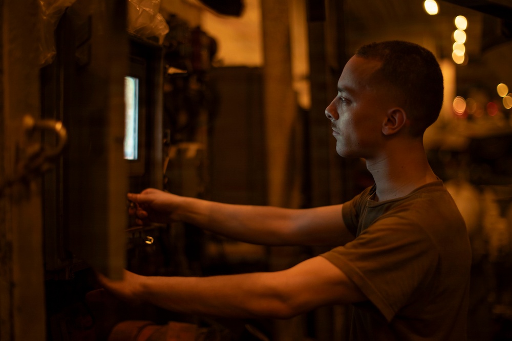 A young man with a short military-style haircut is shown in profile, working at what appears to be an industrial panel or equipment in a dimly lit environment. He is wearing an olive/khaki t-shirt and is reaching forward with both hands to operate or adjust controls. The scene is illuminated by warm amber/orange ambient light with a small blue screen visible on the panel.