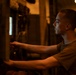 A young man with a short military-style haircut is shown in profile, working at what appears to be an industrial panel or equipment in a dimly lit environment. He is wearing an olive/khaki t-shirt and is reaching forward with both hands to operate or adjust controls. The scene is illuminated by warm amber/orange ambient light with a small blue screen visible on the panel.