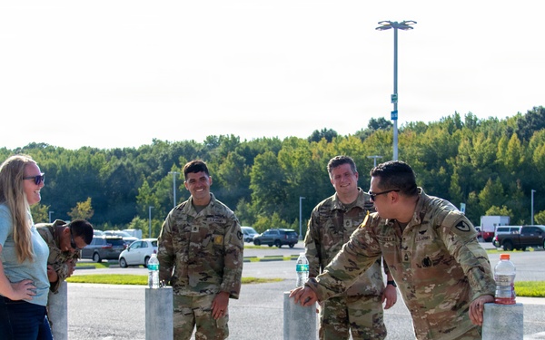 A group of U.S. Army soldiers in OCP (Operational Camouflage Pattern) uniforms are gathered near bollards in a parking lot, conversing with a civilian woman in casual clothing. Military rucksacks are visible on the ground nearby, suggesting the group may have recently completed or is preparing for a ruck march or physical training event. The setting appears to be a military installation or base facility during daytime.
