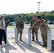 A group of U.S. Army soldiers in OCP (Operational Camouflage Pattern) uniforms are gathered near bollards in a parking lot, conversing with a civilian woman in casual clothing. Military rucksacks are visible on the ground nearby, suggesting the group may have recently completed or is preparing for a ruck march or physical training event. The setting appears to be a military installation or base facility during daytime.