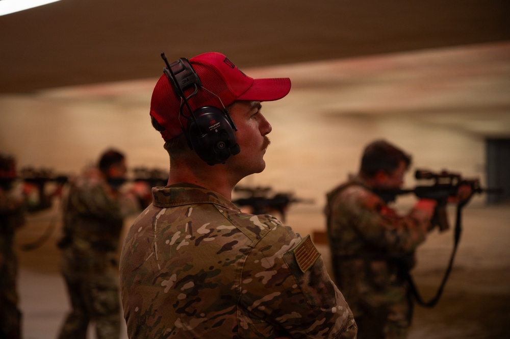 The image shows multiple individuals in military camouflage uniforms at what appears to be an indoor shooting range. The foreground subject wears a red baseball cap and black hearing protection (ear muffs), and is not actively shooting, while others in the background are aiming rifles downrange. The setting appears to be a covered firing range with a sandy or concrete backdrop.