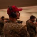 The image shows multiple individuals in military camouflage uniforms at what appears to be an indoor shooting range. The foreground subject wears a red baseball cap and black hearing protection (ear muffs), and is not actively shooting, while others in the background are aiming rifles downrange. The setting appears to be a covered firing range with a sandy or concrete backdrop.