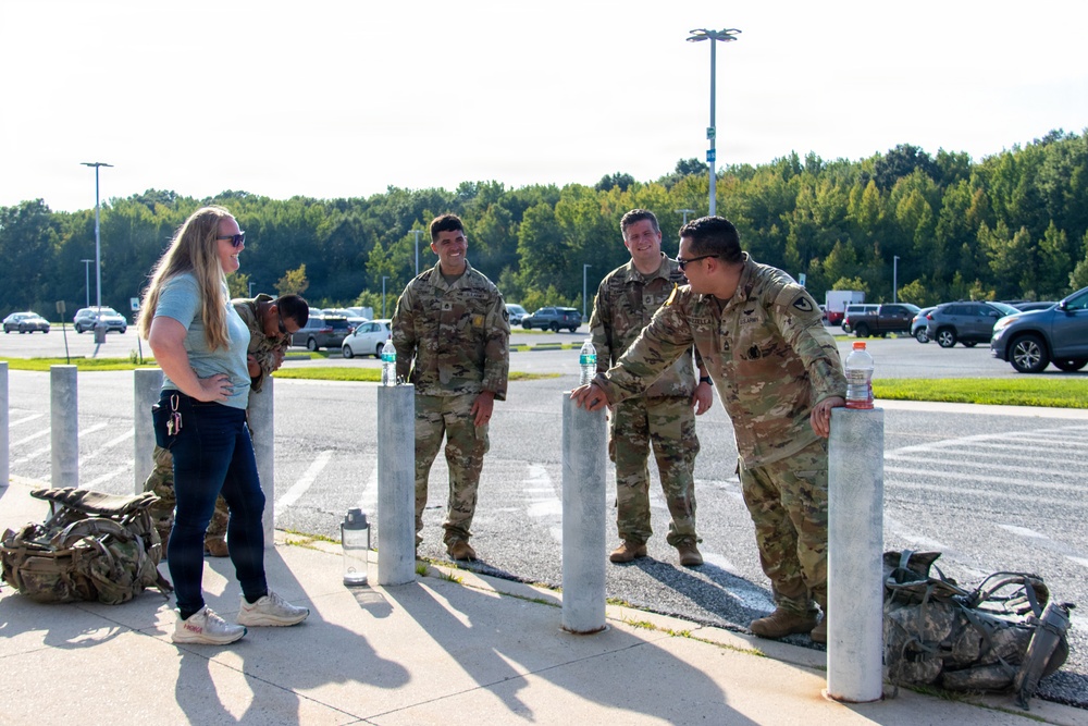 A group of U.S. Army soldiers in OCP (Operational Camouflage Pattern) uniforms are gathered near bollards in what appears to be a military installation parking lot. They are interacting with a civilian woman in casual clothing, appearing relaxed and engaged in conversation. Military rucksacks are visible on the ground nearby, suggesting a recent or upcoming ruck march or physical training event.