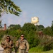 Two U.S. Army soldiers in OCP (Operational Camouflage Pattern) uniforms are conducting a ruck march along a paved pathway on what appears to be a military installation. Both soldiers are wearing full rucksacks and sunglasses, with one carrying a red water bottle. A military water tower with an insignia is visible in the background.