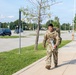 A smiling soldier in US Army Operational Camouflage Pattern (OCP) uniform walks along a sidewalk on what appears to be a military installation. He is carrying a large rucksack on his back and holding a water bottle, suggesting he is completing or participating in a ruck march. The setting includes a large parking lot, lamp posts, trees, and well-maintained grounds consistent with a military base.