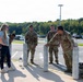 A group of military personnel in OCP (Operational Camouflage Pattern) uniforms are gathered near concrete bollards in what appears to be a military installation parking lot. They are interacting with a civilian woman in casual clothing. Military rucksacks are visible on the ground nearby, suggesting a ruck march or physical training event.