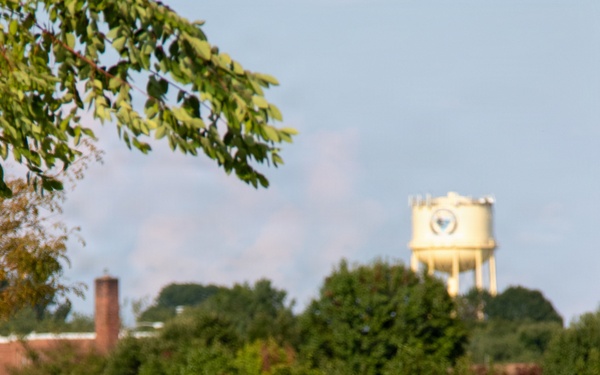 Two U.S. Army soldiers in Operational Camouflage Pattern (OCP) uniforms are walking along a paved path while carrying rucksacks, consistent with a ruck march or physical training exercise. Both wear sunglasses and have American flag patches on their right shoulders. The setting appears to be a military installation, evidenced by a water tower visible in the background.