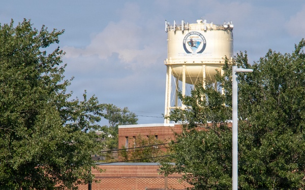 The image shows a military installation or government facility featuring a brick building and a prominent water tower bearing an official seal or emblem. A soldier in camouflage uniform carrying gear is walking across a well-maintained grassy area in the foreground. A dark SUV is parked near the building entrance.