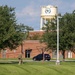 The image shows a military installation or government facility featuring a brick building and a prominent water tower bearing an official seal or emblem. A soldier in camouflage uniform carrying gear is walking across a well-maintained grassy area in the foreground. A dark SUV is parked near the building entrance.