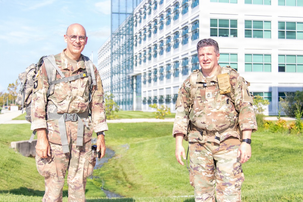 Two U.S. Army soldiers in OCP (Operational Camouflage Pattern) uniforms are walking outdoors near a modern glass office building. Both are wearing rucksacks and appear to be in good spirits, smiling at the camera. The setting suggests a military installation or government campus.
