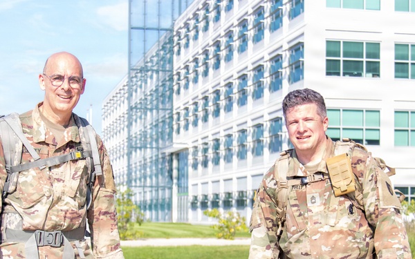 Two military personnel in U.S. Army OCP (Operational Camouflage Pattern) uniforms are posing outdoors in front of a large modern glass-and-steel building. Both are wearing rucksacks and tactical gear, suggesting they are participating in a ruck march or physical training event. The setting appears to be a military installation or government campus on a clear, sunny day.