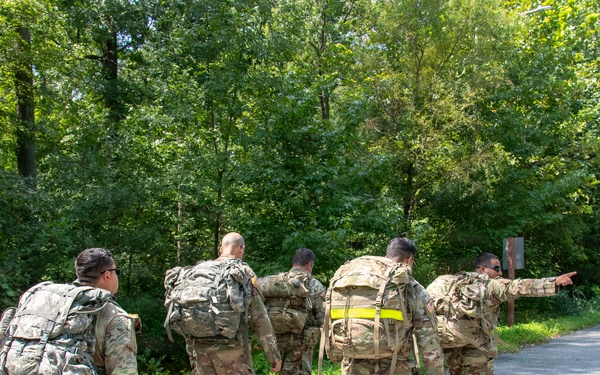 A group of five military personnel in camouflage uniforms are conducting a road march along a paved road bordered by dense green forest. They are carrying large rucksacks on their backs, and one individual at the front right is pointing in a direction, likely providing navigation guidance. The scene appears to be a training exercise or ruck march event during daylight hours.