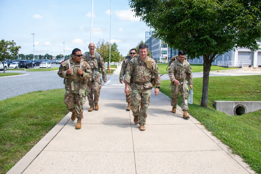 A group of five military personnel in U.S. Army OCP (Operational Camouflage Pattern) uniforms are walking along a sidewalk on what appears to be a military installation. They are carrying rucksacks and tactical gear, suggesting they are conducting or completing a ruck march or physical training exercise. The setting includes well-maintained grounds, modern buildings, and a parking area consistent with a U.S. Army base.