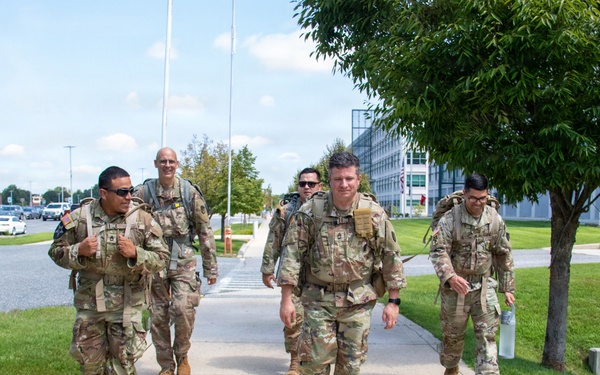 A group of five military personnel in U.S. Army OCP (Operational Camouflage Pattern) uniforms are walking along a sidewalk on what appears to be a military installation. They are carrying rucksacks and tactical gear, suggesting they are conducting or completing a ruck march or physical training exercise. The setting includes well-maintained grounds, modern buildings, and a parking area consistent with a U.S. Army base.