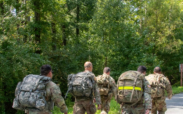 A group of five military personnel in camouflage uniforms are conducting a road march along a paved road bordered by dense green vegetation. They are all carrying large rucksacks and wearing standard-issue combat boots, moving away from the camera. One soldier has a high-visibility yellow reflective band on their pack, likely for safety identification.