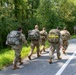 A group of five military personnel in camouflage uniforms are conducting a road march along a paved road bordered by dense green vegetation. They are all carrying large rucksacks and wearing standard-issue combat boots, moving away from the camera. One soldier has a high-visibility yellow reflective band on their pack, likely for safety identification.