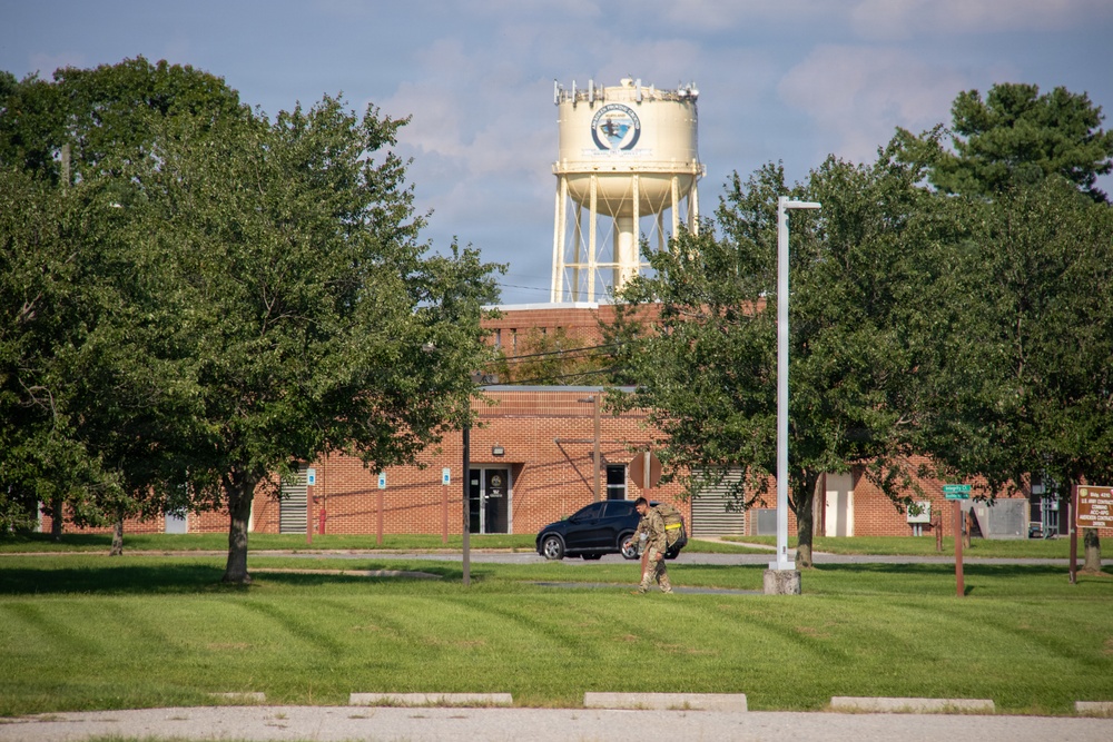 The image shows what appears to be a U.S. military installation or government facility, featuring a brick building, a prominent water tower with an emblem or seal, and well-maintained grounds. A person in military camouflage uniform is walking near the building, with a dark SUV parked nearby. The scene is set during daytime with lush green trees and a partly cloudy sky.