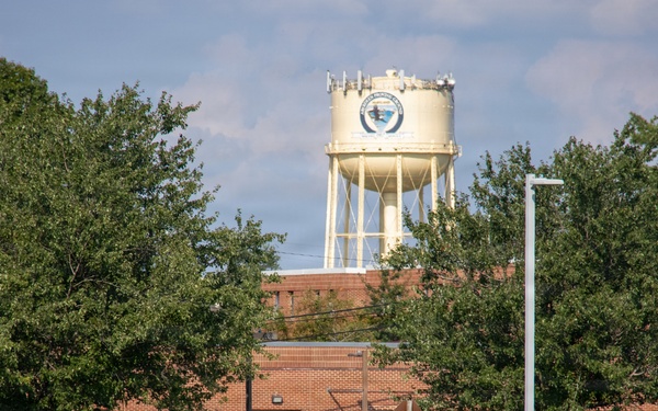 The image shows what appears to be a U.S. military installation or government facility, featuring a brick building, a prominent water tower with an emblem or seal, and well-maintained grounds. A person in military camouflage uniform is walking near the building, with a dark SUV parked nearby. The scene is set during daytime with lush green trees and a partly cloudy sky.