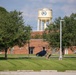 The image shows what appears to be a U.S. military installation or government facility, featuring a brick building, a prominent water tower with an emblem or seal, and well-maintained grounds. A person in military camouflage uniform is walking near the building, with a dark SUV parked nearby. The scene is set during daytime with lush green trees and a partly cloudy sky.