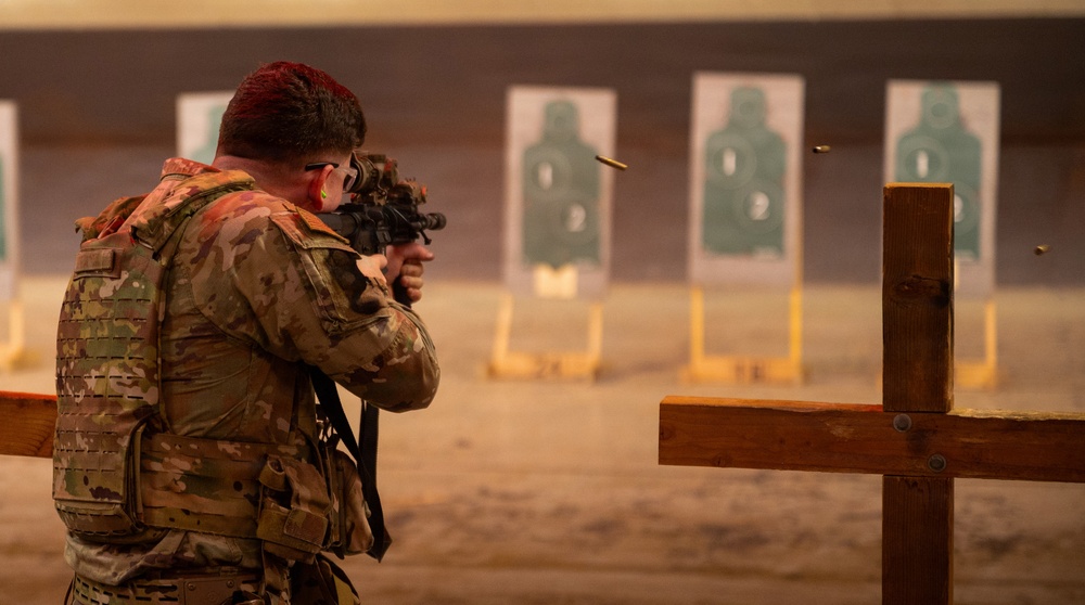 A soldier in full multicam camouflage uniform and tactical vest is firing a rifle at a shooting range. Multiple paper silhouette targets are visible downrange, and a spent brass casing can be seen ejecting from the weapon. The shooter is positioned behind a wooden barricade structure.