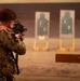 A soldier in full multicam camouflage uniform and tactical vest is firing a rifle at a shooting range. Multiple paper silhouette targets are visible downrange, and a spent brass casing can be seen ejecting from the weapon. The shooter is positioned behind a wooden barricade structure.