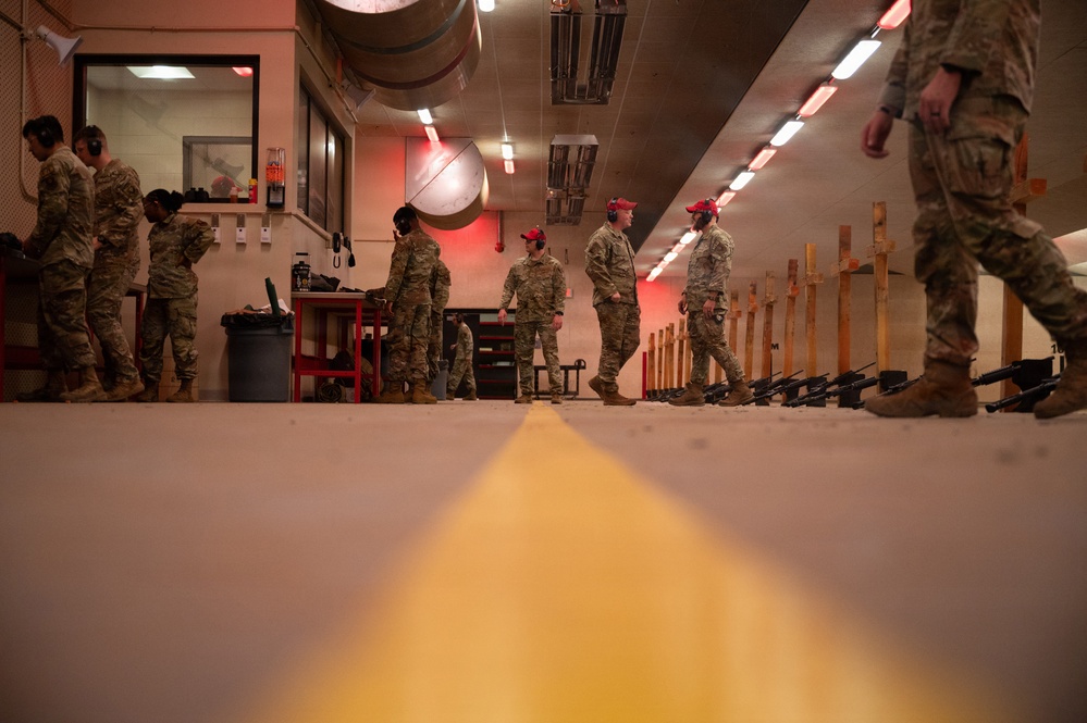 Military personnel in camouflage uniforms are gathered inside an indoor shooting range. Several individuals wearing red caps, likely range safety officers or instructors, are visible among the group. Rifles are laid out on the floor along the firing line, and a yellow safety line is prominently visible in the foreground.